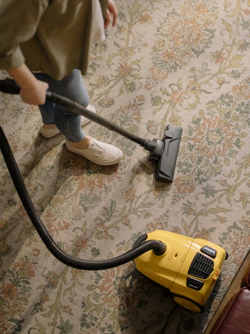 A person vacuuming a traditional patterned area rug with a yellow canister vacuum cleaner in a well-lit room. The individual is wearing beige shoes, blue jeans, and a brown jacket. The vacuum's black