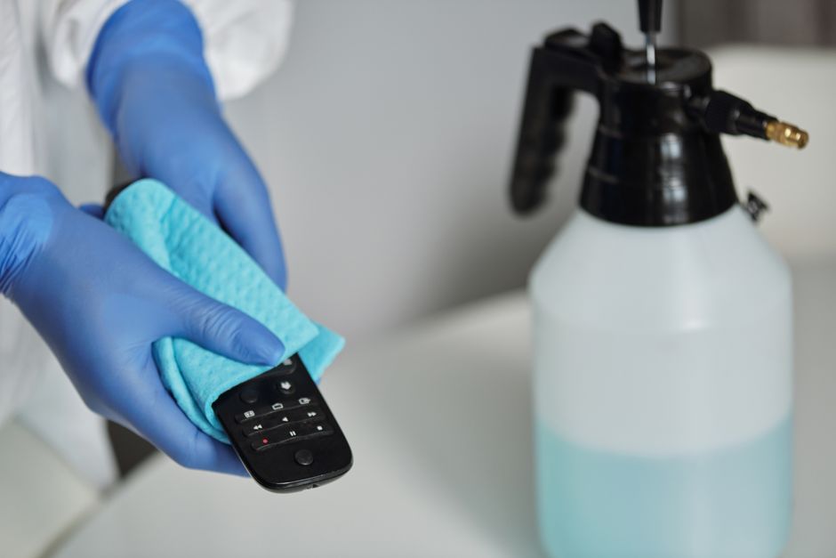 A person wearing black gloves using a yellow handheld upholstery cleaning machine to treat a stain on a dark grey fabric sofa in a residential living room. The sofa has a modern design with padded arm