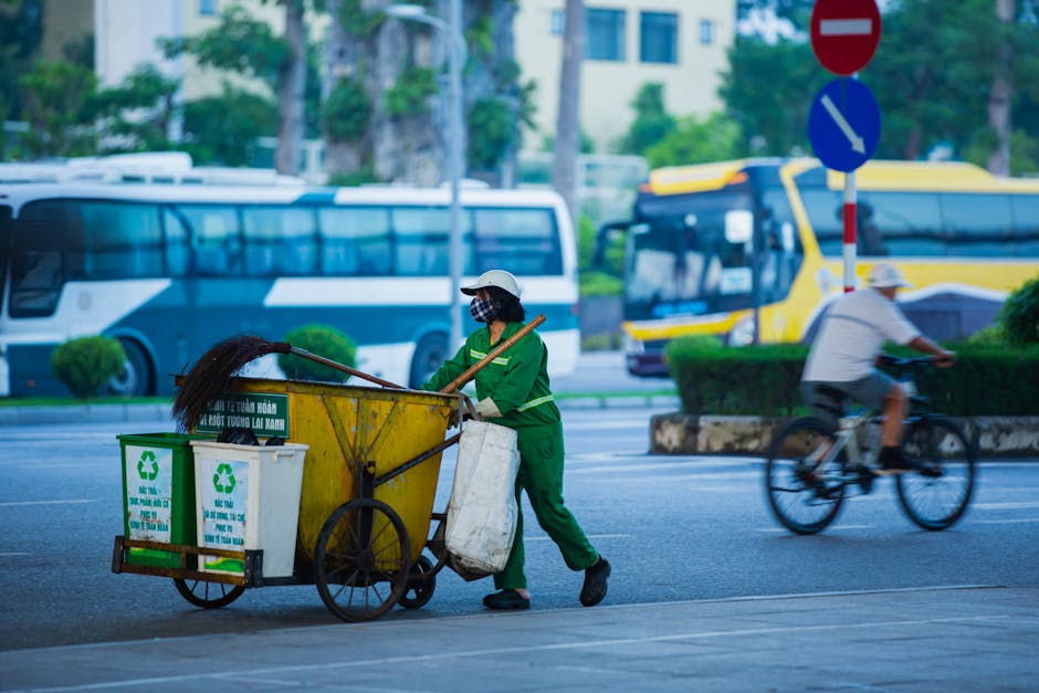 A street scene showing a sanitation worker in a green uniform and protective face mask, pushing a yellow wheeled trash cart with recycling bins attached, while a cyclist wearing a white t-shirt and he