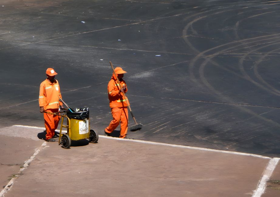 A street scene showing a sanitation worker in a green uniform and protective face mask, pushing a yellow wheeled trash cart with recycling bins attached, while a cyclist wearing a white t-shirt and he